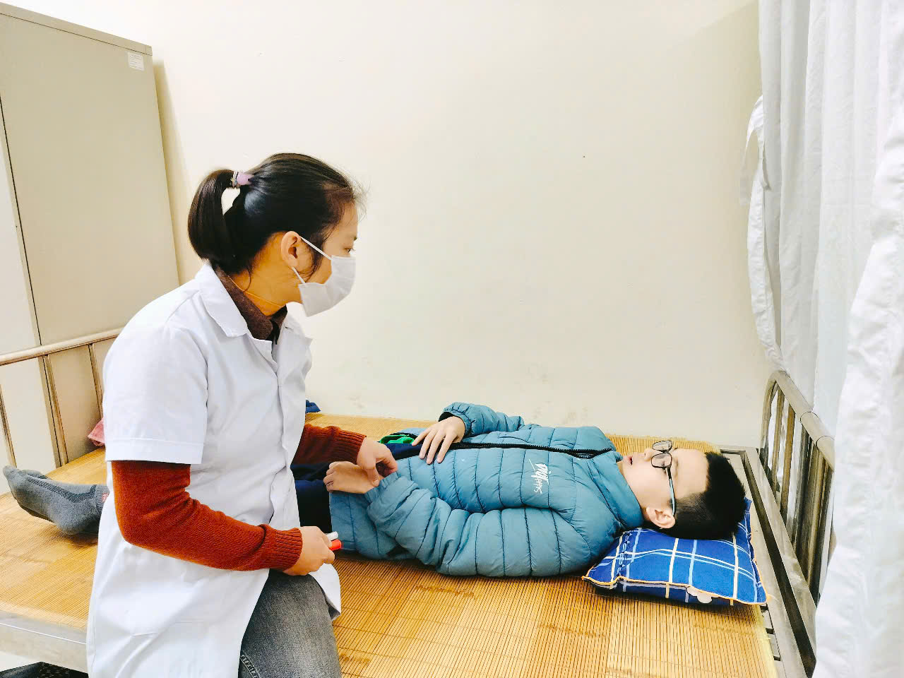 A person in a white coat and mask examining a child lying on a bedDescription automatically generated