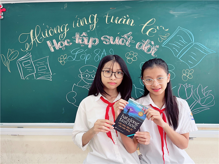 Two girls in white shirts and red ties holding a book in front of a chalkboardDescription automatically generated