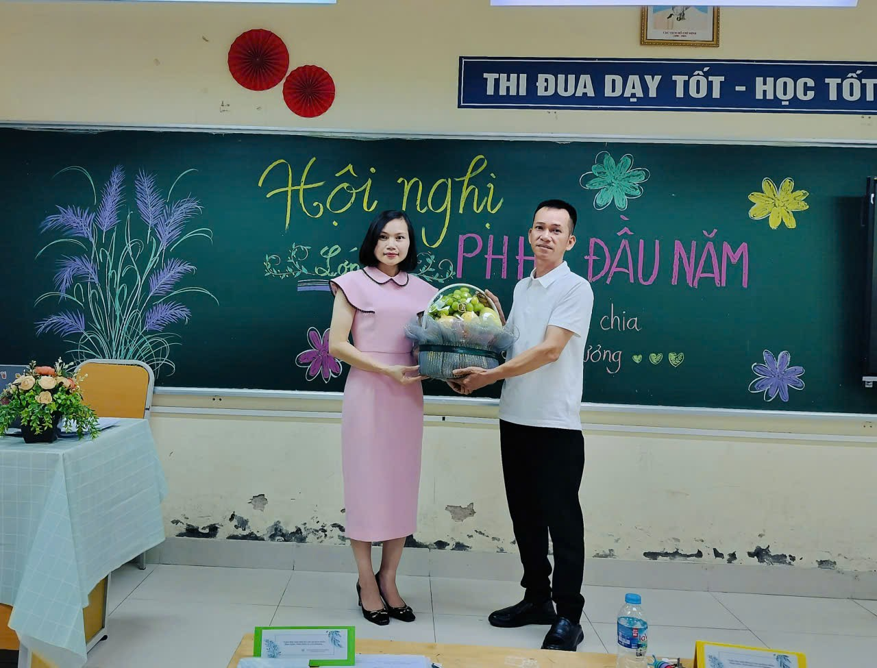 A person and person holding a basket of fruit in front of a chalkboard
Description automatically generated
